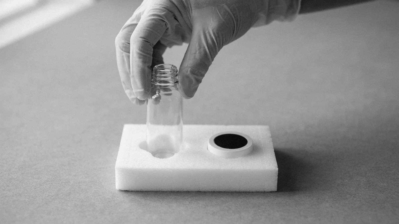 A gloved hand lifts a clear glass bottle from a foam insert beside a round white monitoring device.