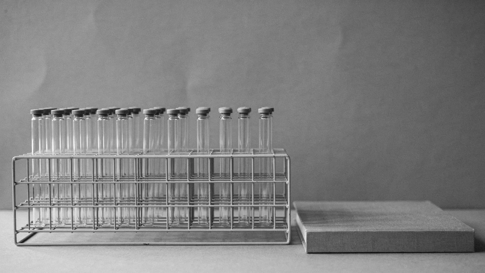 A wire rack holding rows of capped clear glass tubes beside a closed cloth-covered binder on a bench.