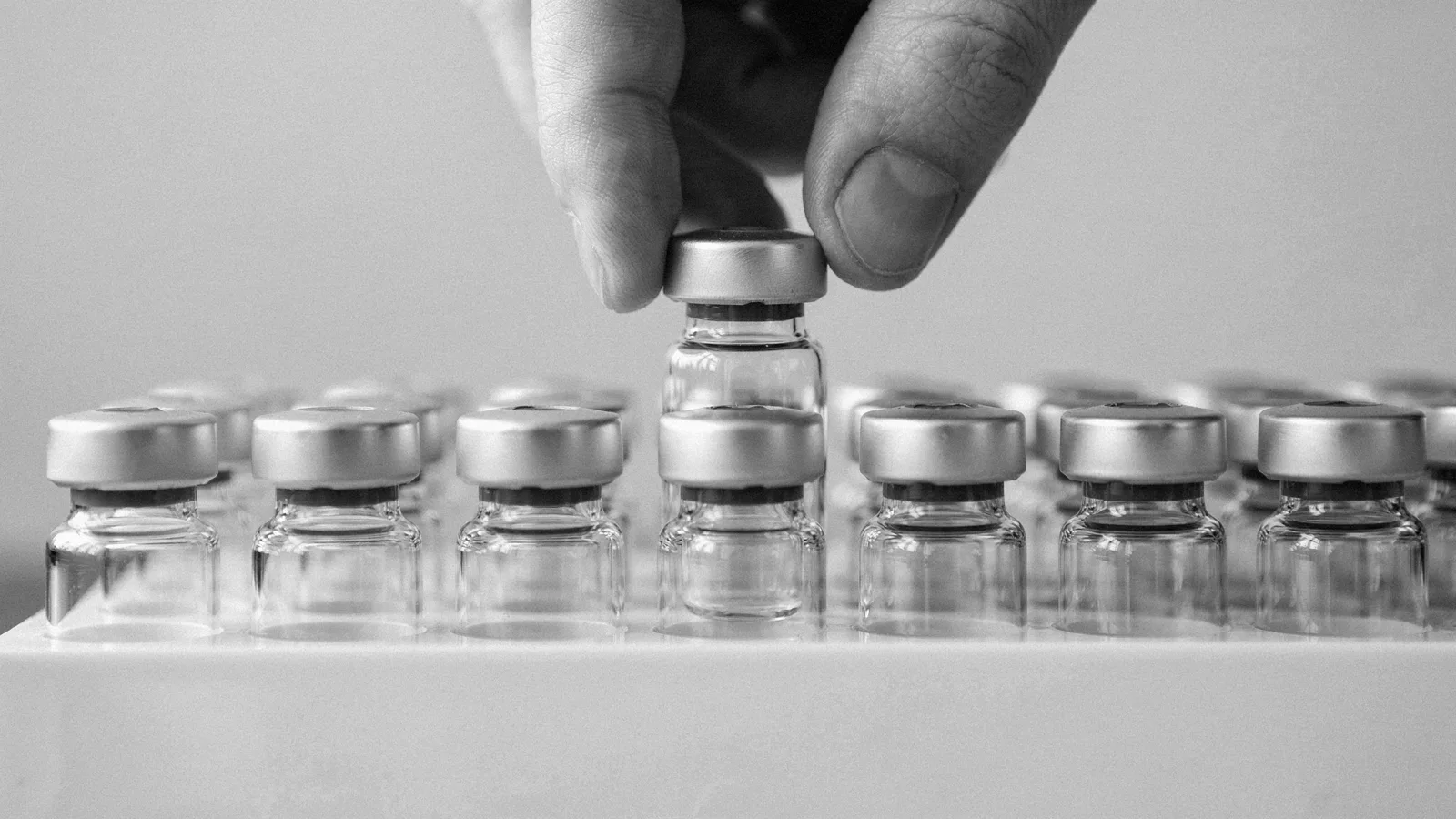Fingers lowering a small clear capped glass vial into a white rack filled with identical vials