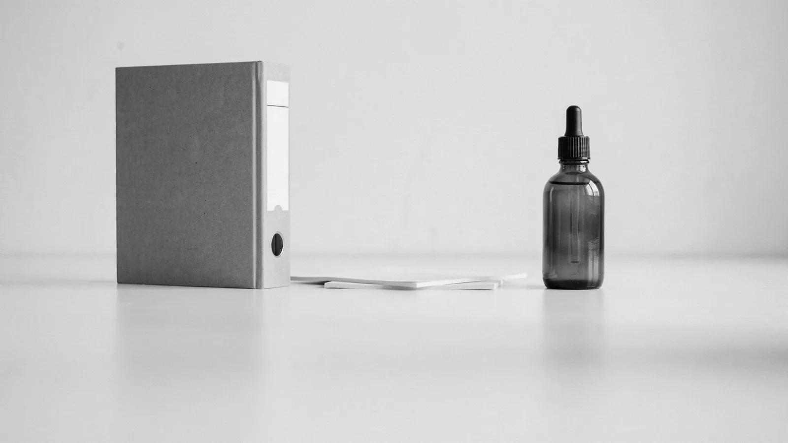 A closed gray binder, a few blank sheets of paper and an amber glass dropper bottle on a white bench