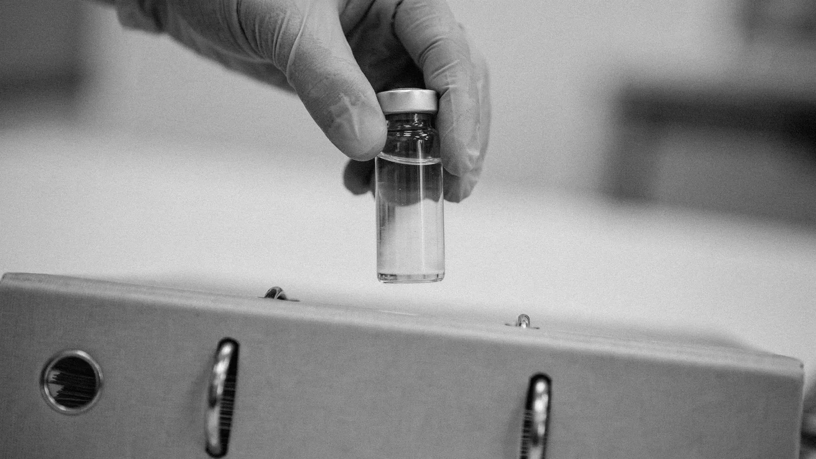 Gloved hands holding a small bare glass vial above a closed plain ring binder on a bench