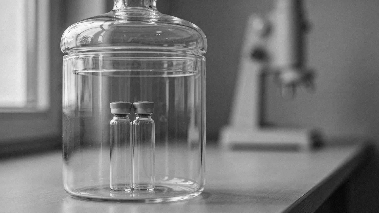 Two small capped glass vials inside a closed glass desiccator, with a lab instrument blurred behind