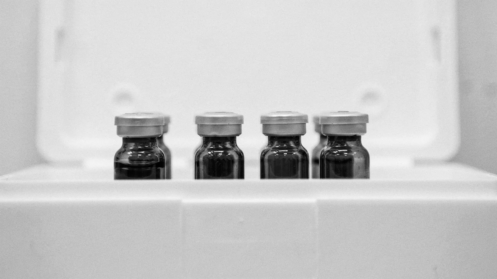 Four small amber glass vials with metal caps standing in a row inside an open white storage box