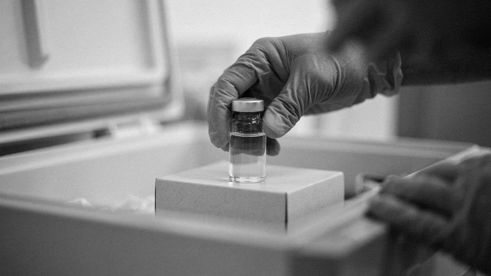 Gloved hand setting a small clear glass vial with a metal cap onto a white box inside an open freezer