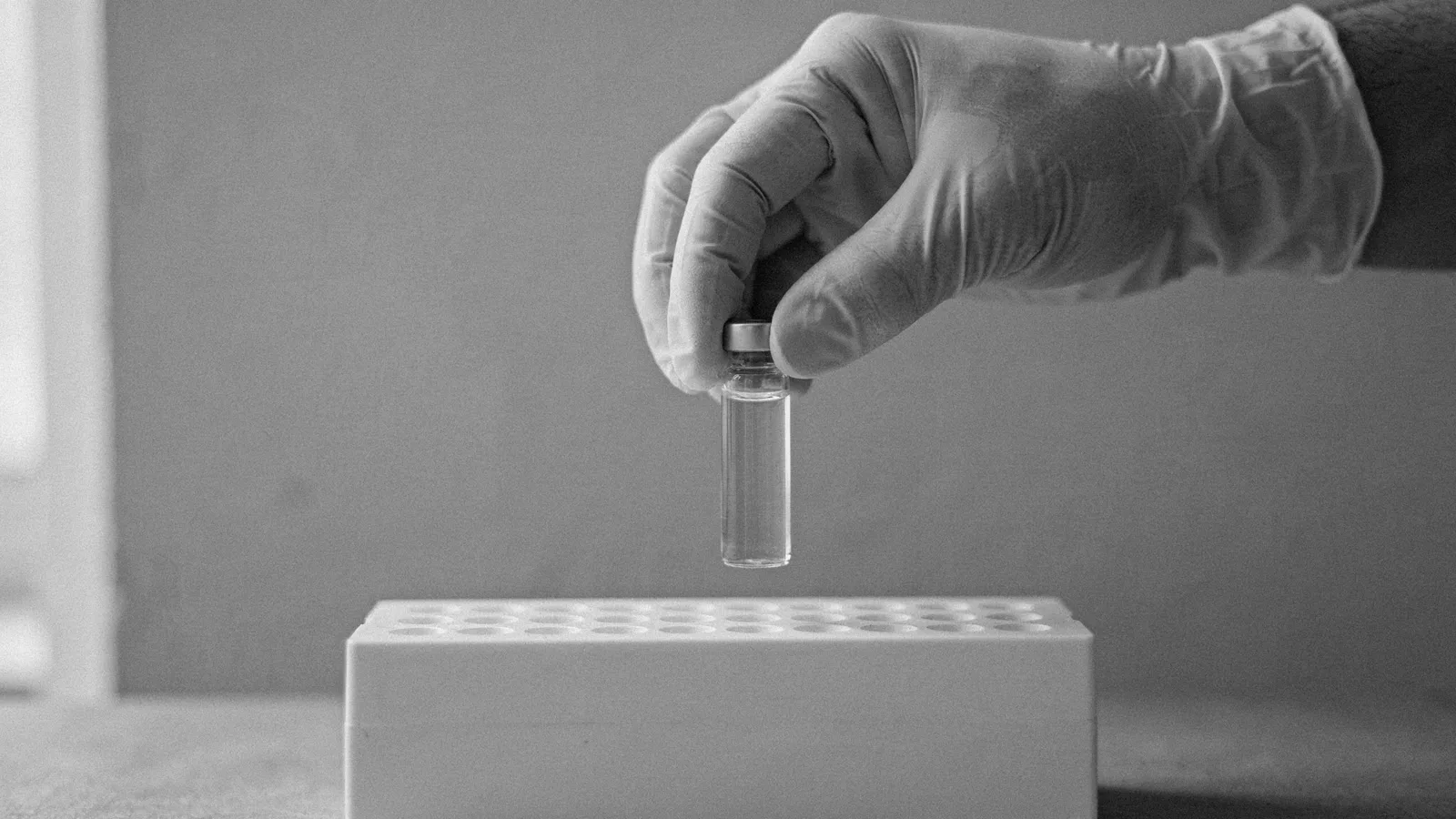 A gloved hand holding a small clear glass vial above a white sample rack in soft light