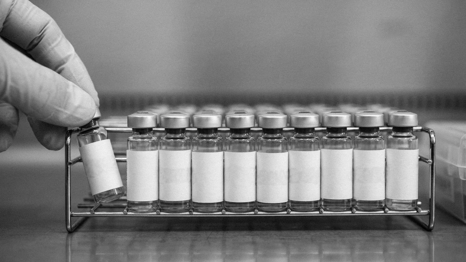 A gloved hand lifting one blank-labelled vial from a wire rack of identical vials