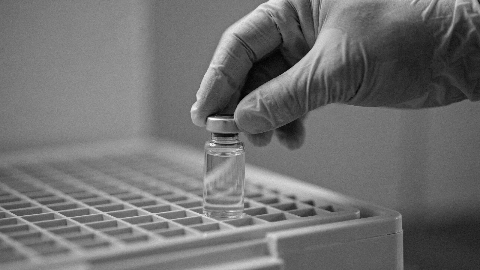 Gloved hands placing a small glass vial into a freezer rack on a laboratory bench