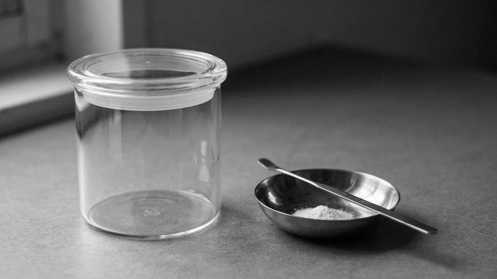 A glass desiccator on a lab bench beside a small metal weighing boat of white powder and a spatula