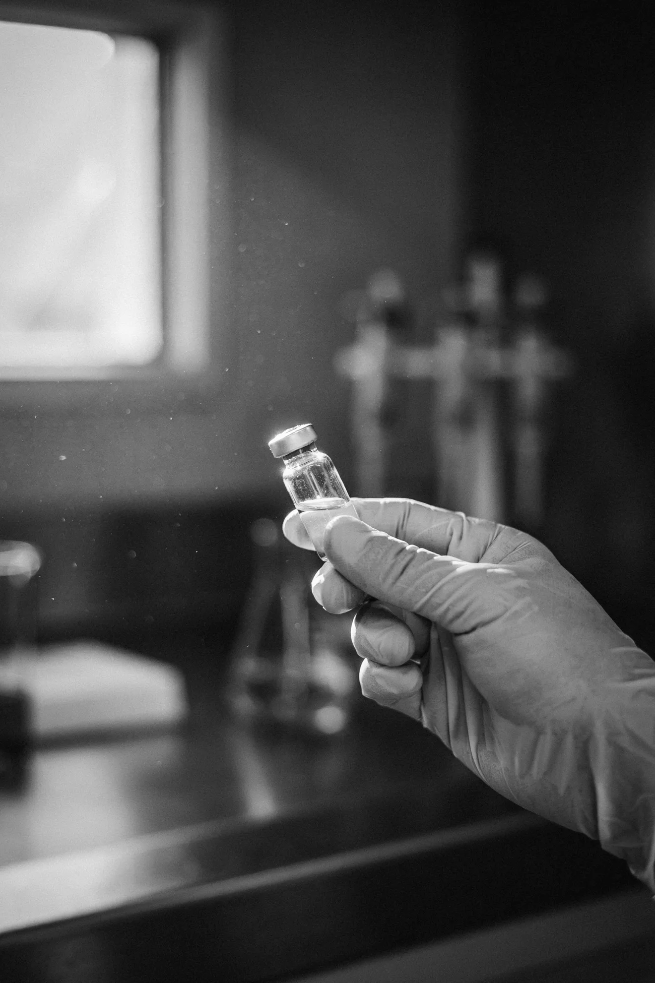 A gloved hand holding a small crimp-capped vial up to a window, in black and white