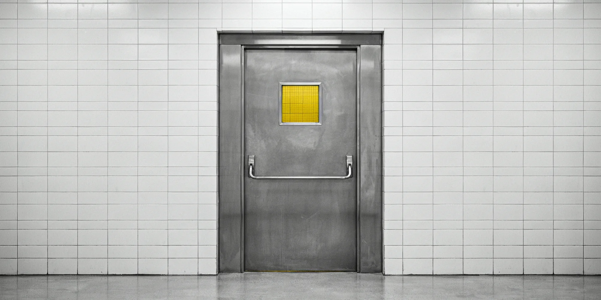 A steel laboratory pass-through door with a small yellow-tinted wired-glass window, set in a white tiled wall